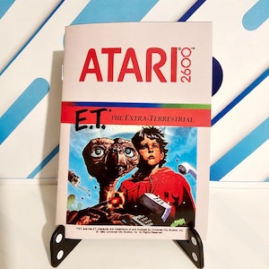 Puede incluir: Un cartucho de juego Atari 2600 vintage para E.T. El Extraterrestre. El cartucho presenta una imagen colorida de E.T. y un niño.