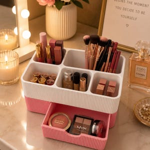 Peut inclure: Un organisateur de maquillage blanc et rose à deux niveaux rempli de divers cosmétiques, notamment des rouges à lèvres, des pinceaux et des palettes de fards à paupières. Le tiroir est ouvert, révélant d'autres produits de maquillage. Une affiche encadrée dit "BEAUTY BEGINS THE MOMENT YOU DECIDE TO BE YOURSELF."