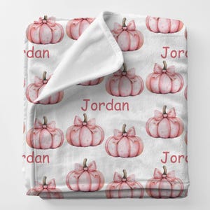 May include: A white fleece blanket featuring a pattern of pink pumpkins with brown stems and pink bows. The name "Jordan" is printed in red across the blanket. The blanket is folded over, revealing a white interior.