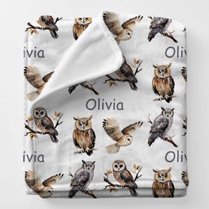 May include: A white blanket featuring a repeating pattern of watercolor owls in various poses, perched on branches or in flight. The name "Olivia" is printed in gray. The blanket is soft and cozy.