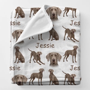 May include: White blanket featuring a repeating pattern of Weimaraner dogs in various poses. The blanket also has the name "Jessie" printed in brown. The dogs are a gray color.