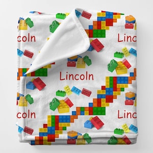 May include: A white blanket featuring a colorful pattern of toy building blocks in red, green, yellow, and blue. The name "Lincoln" is printed in red across the blanket.