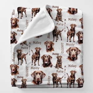 Puede incluir: Una manta blanca con un patr&oacute;n repetido de perros Labrador Retriever marrones en varias poses. La palabra "Maisy" est&aacute; impresa verticalmente junto a las ilustraciones de los perros. La manta est&aacute; doblada, revelando un reverso blanco.
