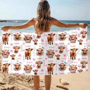 Puede incluir: Toalla de playa blanca con un patr&oacute;n repetido de vacas de las Tierras Altas marrones, arreglos florales y flores rosas. El nombre "Isabel" est&aacute; impreso en la toalla. La toalla se sostiene en una playa.