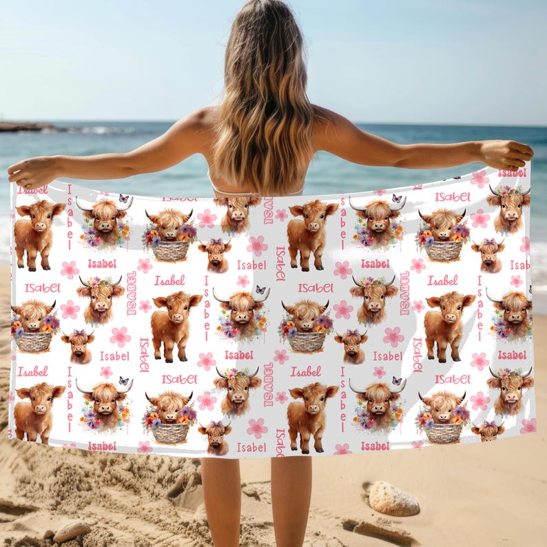 Puede incluir: Toalla de playa blanca con un patr&oacute;n repetido de vacas de las Tierras Altas marrones, arreglos florales y flores rosas. El nombre "Isabel" est&aacute; impreso en la toalla. La toalla se sostiene en una playa.