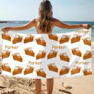 May include: White beach towel featuring a repeating pattern of pumpkin pie slices with whipped cream. The word "Parker" is printed in brown. The towel is held up on a beach with the ocean in the background.