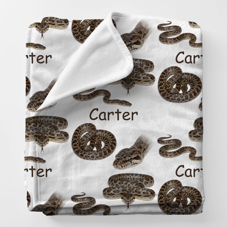 May include: White blanket with a repeating pattern of brown and tan snakes in different positions. The word "Carter" is printed in brown on the blanket. The blanket is folded over, showing a white interior.