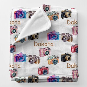 May include: White fleece blanket with a repeating pattern of watercolor cameras in various colors, including pink, blue, and orange. The name "Dakota" is printed in brown on the blanket.