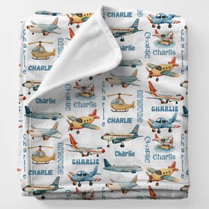 May include: White baby blanket featuring a pattern of various airplanes in shades of blue, yellow, and red. The name "Charlie" is printed in blue throughout the design. The blanket is soft and cozy.