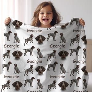 K&ouml;nnte beinhalten: Wei&szlig;e Decke mit sich wiederholendem Muster von Deutsch Kurzhaar Hunden und dem Namen "Georgie". Die Hunde sind braun und grau. Die Decke wird von einem l&auml;chelnden Kind gehalten. Ein gem&uuml;tliches Wohnaccessoire.