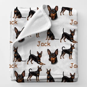 May include: White blanket featuring a repeating pattern of black and brown Miniature Pinscher dogs in various poses. The word "Jack" is printed in brown on the blanket. The blanket is folded over, revealing the white underside.