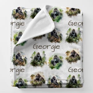 May include: White blanket featuring a repeating pattern of watercolor-style gorilla illustrations. Each gorilla is surrounded by green foliage. The name "Georgie" is printed in a brown script font across the blanket.