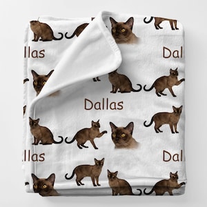 May include: White fleece blanket featuring repeating images of brown cats with yellow eyes. The word "Dallas" is printed in brown. The blanket is folded over, showing the white underside.