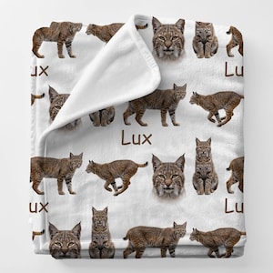 May include: White blanket featuring multiple images of bobcats in various poses. The blanket has a repeating pattern of brown bobcats and the word "Lux" in brown. The blanket is folded over, showing the white underside.