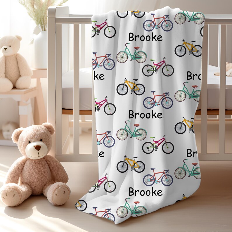 May include: White baby blanket with a repeating pattern of colorful bicycles and the name "Brooke" in black. The bicycles are in shades of red, green, and yellow. The blanket is draped over a crib.