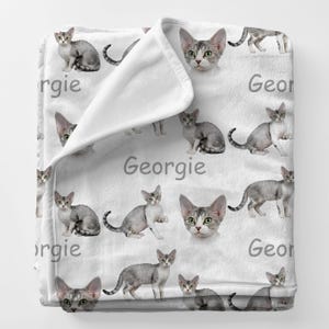 May include: A white blanket featuring a pattern of gray cats and the name "Georgie" in gray text. The cats are in various poses, including sitting and standing. The blanket is folded over, revealing a soft texture.