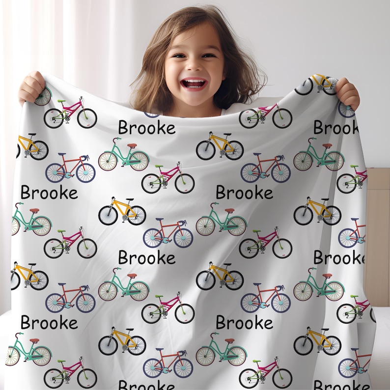May include: White blanket featuring a repeating pattern of colorful bicycles in red, yellow, and teal. The name "Brooke" is printed in black script throughout the design. The blanket is held up by a smiling child.
