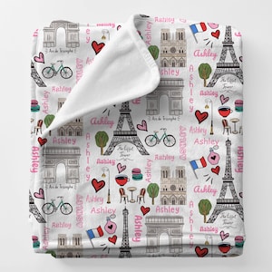 May include: White throw blanket with a repeating pattern of Parisian landmarks, including the Eiffel Tower, Arc de Triomphe, and Notre Dame Cathedral. The design also features bicycles, hearts, and the name "Ashley" in pink script.