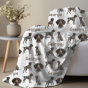 K&ouml;nnte beinhalten: Wei&szlig;e Decke mit einem sich wiederholenden Muster aus Deutsch Kurzhaar und dem Namen "Georgie" in Schreibschrift. Die Decke ist &uuml;ber ein wei&szlig;es Sofa drapiert.