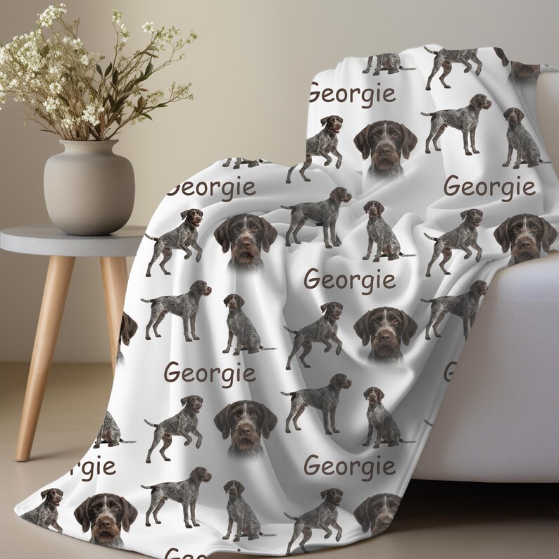 K&ouml;nnte beinhalten: Wei&szlig;e Decke mit einem sich wiederholenden Muster aus Deutsch Kurzhaar und dem Namen "Georgie" in Schreibschrift. Die Decke ist &uuml;ber ein wei&szlig;es Sofa drapiert.