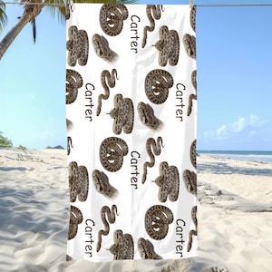 May include: White beach towel with a repeating pattern of brown snakes and the name "Carter" in black. The snakes are in different poses, some coiled and others with their heads raised. The towel is hanging on a clothesline against a beach background.
