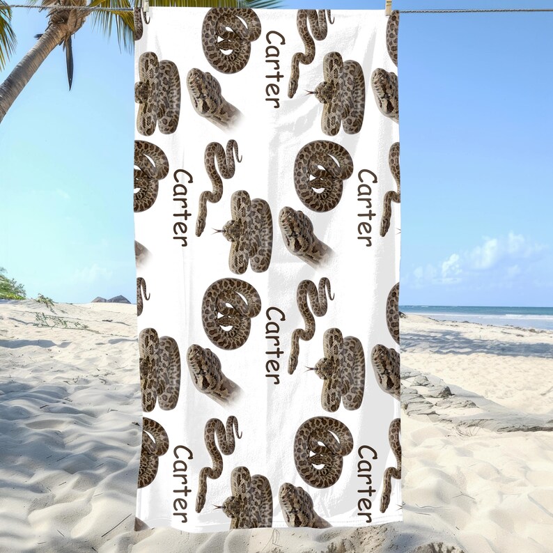 May include: White beach towel with a repeating pattern of brown snakes and the name "Carter" in black. The snakes are in different poses, some coiled and others with their heads raised. The towel is hanging on a clothesline against a beach background.