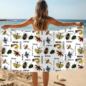 May include: White beach towel featuring a repeating pattern of various insects in shades of brown, green, yellow, and blue. The name "Adrian" is printed in black script throughout the design. The towel is held up on a sandy beach.