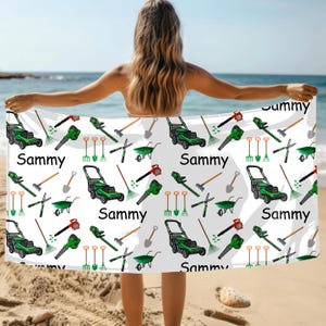 May include: White beach towel with a repeating pattern of green lawn mowers, rakes, shovels, and other gardening tools. The word "Sammy" is printed in black. The towel is held up on a beach with the ocean in the background.