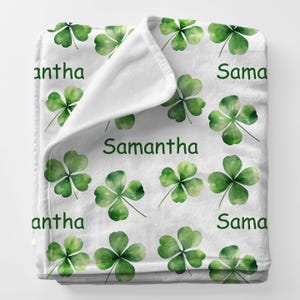 May include: White fleece blanket featuring a pattern of green watercolor shamrocks and the name "Samantha" in green lettering. The blanket is folded over, revealing a soft, plush texture. A cozy and personalized home decor item.