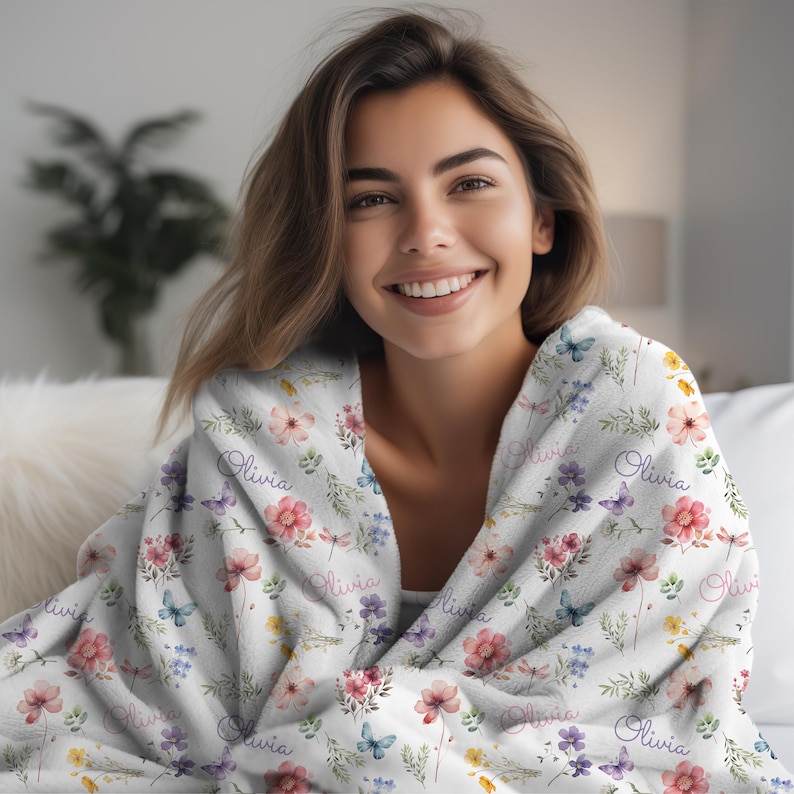 May include: A white floral blanket with the name "Olivia" printed on it. The blanket features a pattern of colorful flowers and butterflies in shades of pink, purple, and yellow. The blanket is draped over a person.