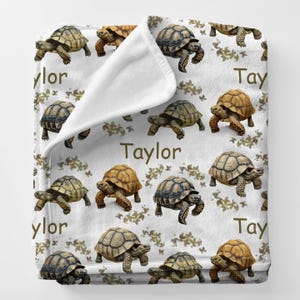 May include: A white blanket featuring a repeating pattern of tortoises in shades of brown and green. The name "Taylor" is printed in brown script across the blanket. The blanket is soft and cozy, perfect for snuggling.