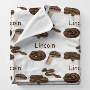 May include: White blanket featuring a repeating pattern of brown snakes in various poses. The word "Lincoln" is printed in brown on the blanket. The blanket is folded over, revealing a white interior.