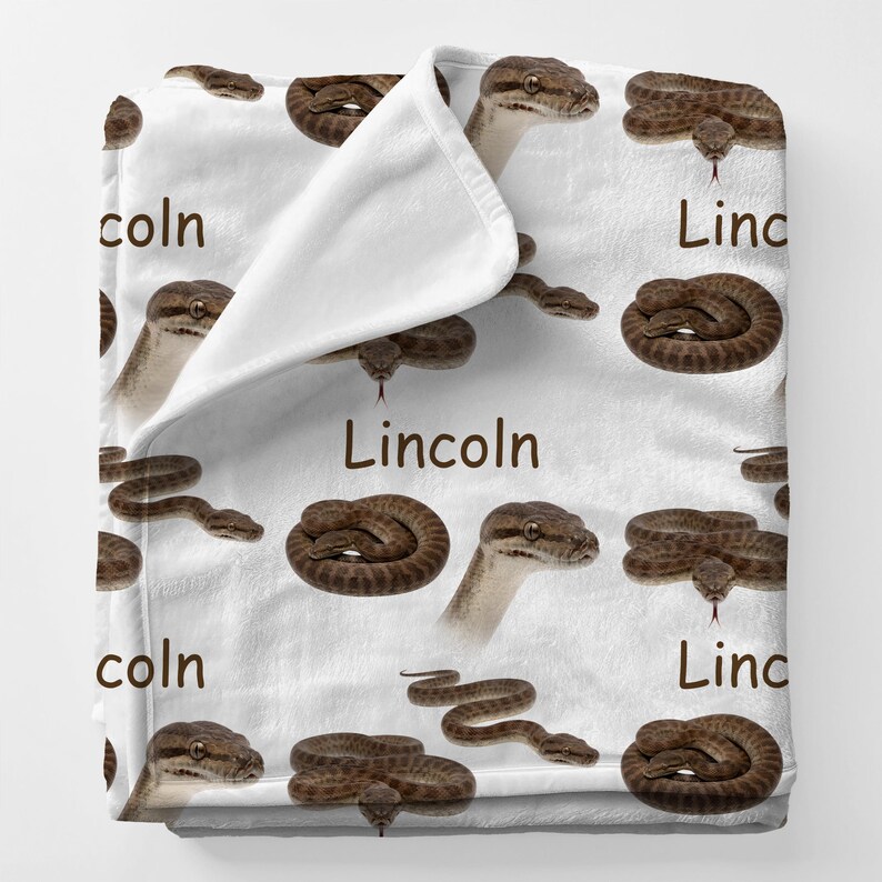 May include: White blanket featuring a repeating pattern of brown snakes in various poses. The word "Lincoln" is printed in brown on the blanket. The blanket is folded over, revealing a white interior.
