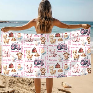 May include: White beach towel featuring a repeating pattern of farm animals, a pink tractor, and a red barn. The name "Emery Ann" is printed in pink. The towel is held up on a beach.