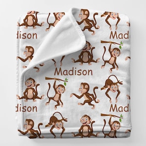 May include: White plush blanket featuring a repeating pattern of cartoon monkeys in various poses. The monkeys are brown with blue eyes and are holding branches. The name "Madison" is printed in white on the blanket.