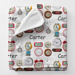 May include: White blanket featuring a repeating pattern of clocks, including alarm clocks, pocket watches, and digital clocks. The name "Carter" is printed in black script across the blanket. The clocks display various times.