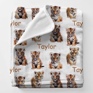 May include: White blanket featuring a repeating pattern of watercolor tiger cubs in shades of orange, brown, and black. The name "Taylor" is printed in brown script across the blanket.