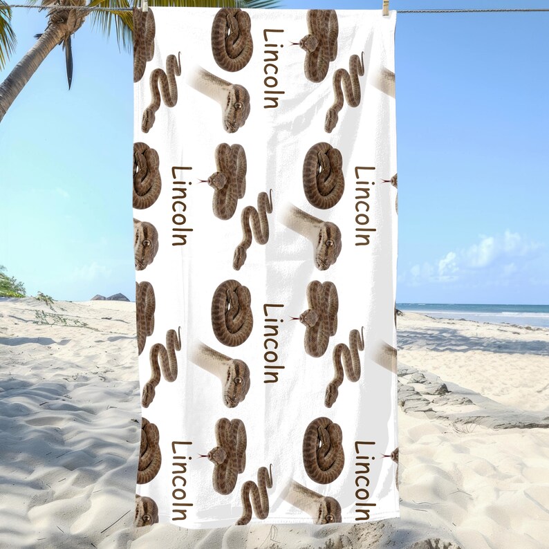 May include: White beach towel with a repeating pattern of brown snakes and the name "Lincoln" printed vertically. The towel is hanging on a clothesline against a beach backdrop.