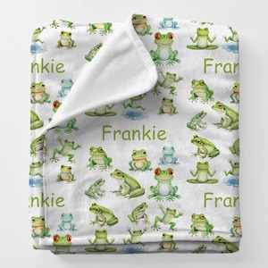 May include: White blanket featuring a pattern of green and blue frogs in various poses. The word "Frankie" is printed in green on the blanket. The blanket is made of a soft, plush material.