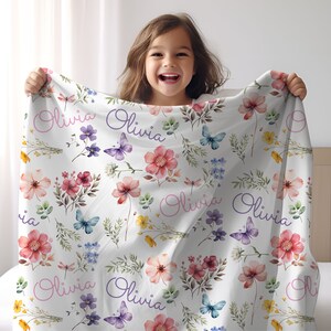 May include: White blanket with a floral pattern featuring pink, red, and yellow flowers, butterflies, and the name "Olivia" in pink and purple script. The blanket is held up by a person.