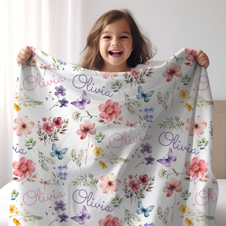 May include: White blanket with a floral pattern featuring pink, red, and yellow flowers, butterflies, and the name "Olivia" in pink and purple script. The blanket is held up by a person.