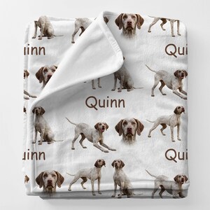 May include: White blanket featuring a repeating pattern of Vizsla dogs in various poses and headshots. The name "Quinn" is printed in brown on the blanket. The blanket is made of a soft, plush material.