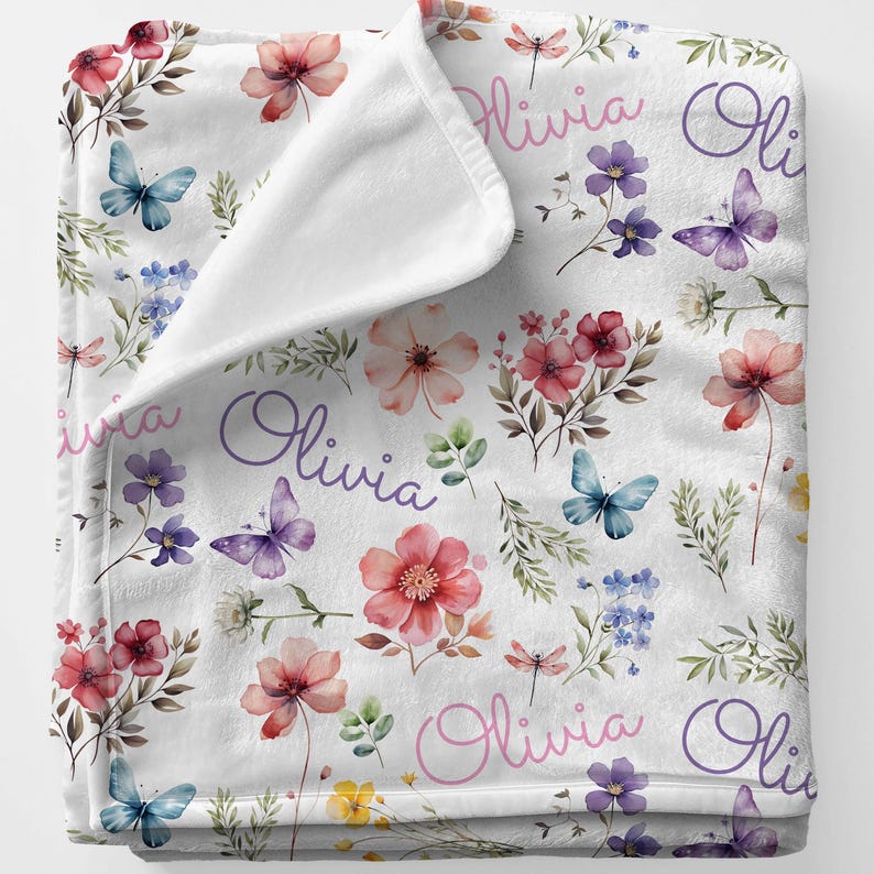 May include: White baby blanket with a floral pattern featuring pink, red, and purple flowers, butterflies, and the name "Olivia" in script. The blanket has a soft, plush texture.