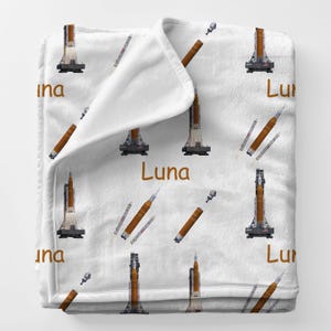 May include: White blanket featuring a repeating pattern of brown and orange rockets and the word "Luna" in brown. The rockets are depicted in various stages of flight, with some appearing to be launching.