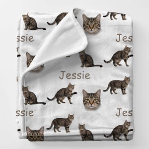 May include: White fleece blanket featuring a repeating pattern of brown tabby cats and the name "Jessie" in brown text. The cats are in various poses, including sitting and standing.