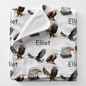 May include: White fleece blanket featuring a repeating pattern of eagles in various poses and colors, including brown, black, and white. The name "Elliot" is printed in black across the blanket.