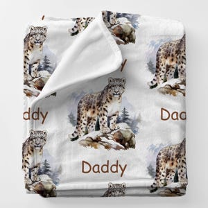 May include: White fleece blanket featuring a repeating pattern of snow leopards in a snowy mountain scene. The words "Daddy" are printed in brown. The blanket is soft and cozy, perfect for snuggling.