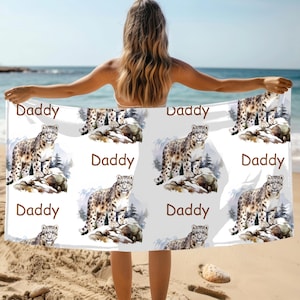 May include: White beach towel featuring a repeating pattern of a snow leopard illustration and the word "Daddy" in brown. The snow leopard is depicted on a snowy rock with a mountain backdrop. The towel is held by a person on a beach.