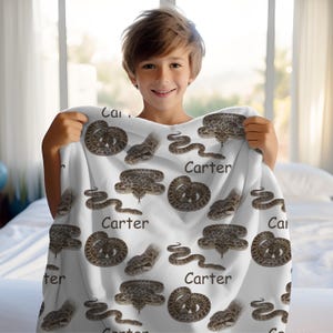 May include: White blanket with a repeating pattern of brown snakes and the name "Carter" in black. The snakes are shown in different poses, including coiled and slithering. The blanket is held up by a person.