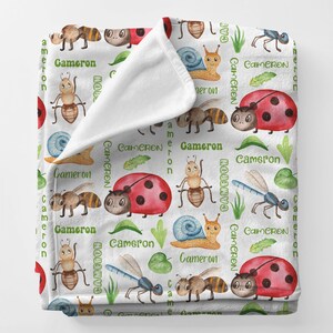 May include: White blanket with a repeating pattern of watercolor insects, including ants, ladybugs, snails, and dragonflies. The name "Cameron" is printed in green. The blanket is folded over, revealing a white interior.